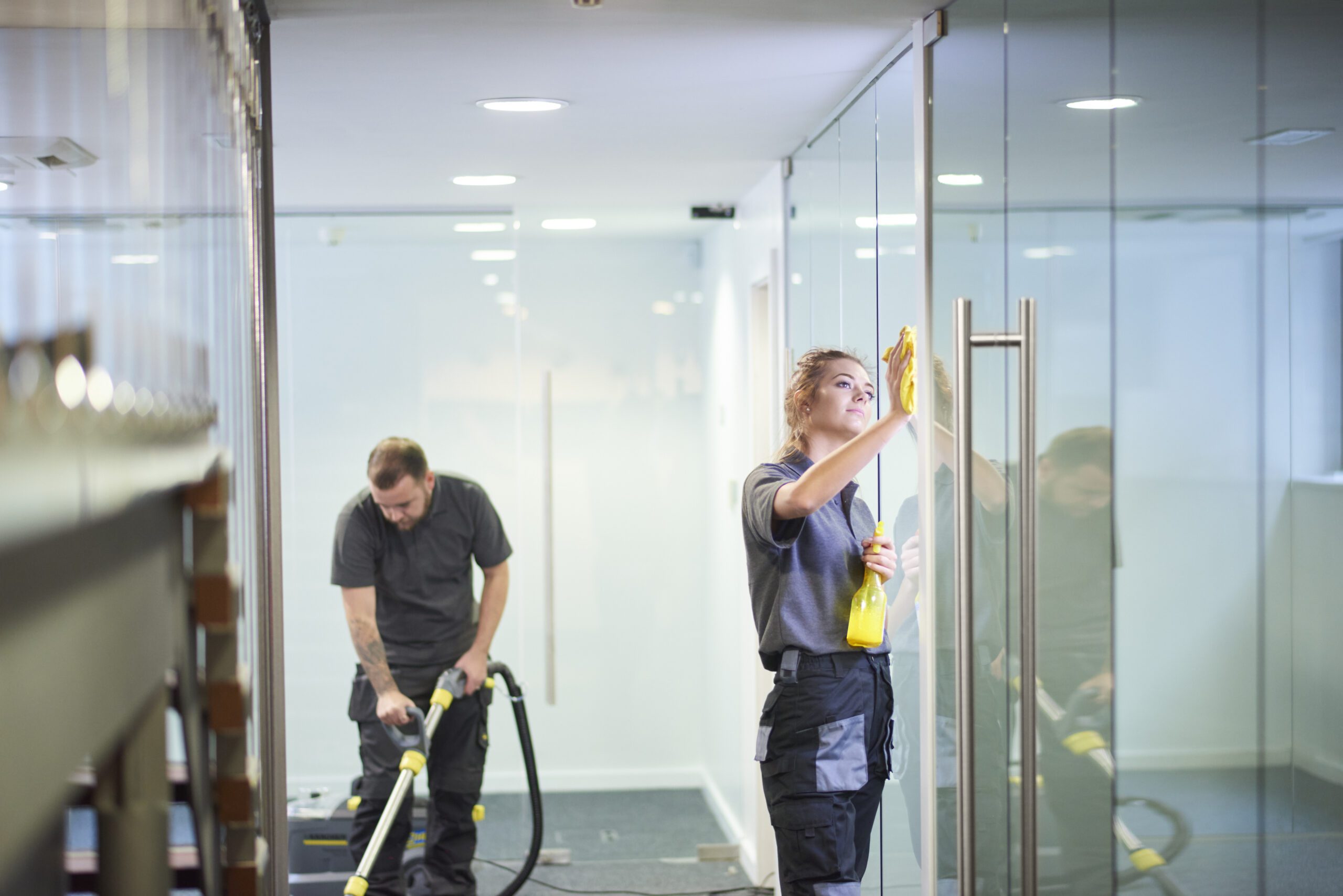 a female cleaning contractor is polishing the glass partition offices whilst In the background a male colleague steam cleans an office carpet in a empty office in between tenants. .